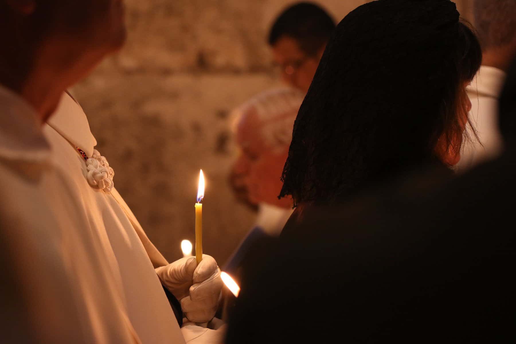 A woman holds a lit, tiny taper candle to her chest in a vigil.