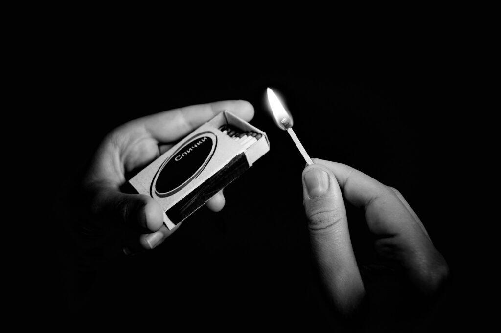 This black and white image shows a close up of two hands holding a matchbook and a lit match.