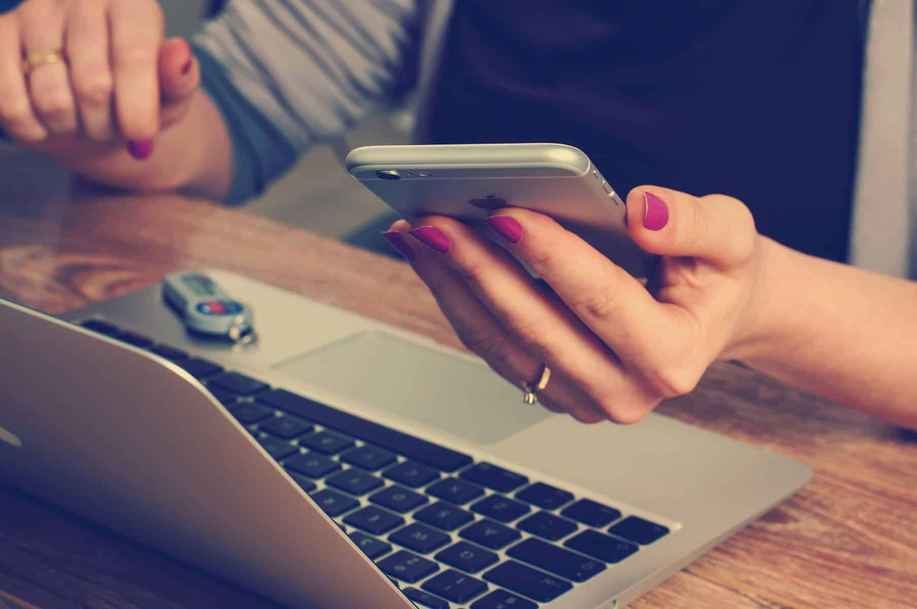 A woman's hands are shown holding a cell phone over a laptop at a desk.