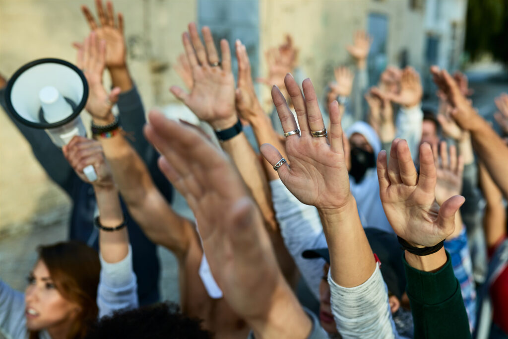 The hands of many people are raised in the air in an outside setting, one holding a bullhorn.