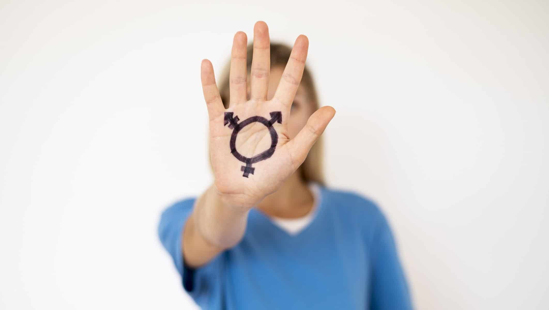 A person stands behind their forward thrust hand with the transgender sign marked on it.