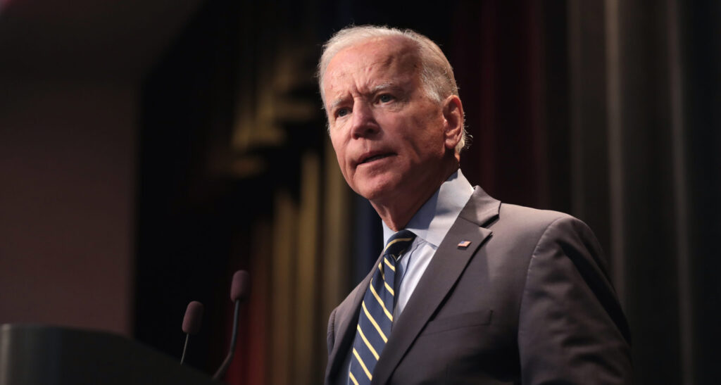 President Biden is shown standing in front of a black curtain and speaking.