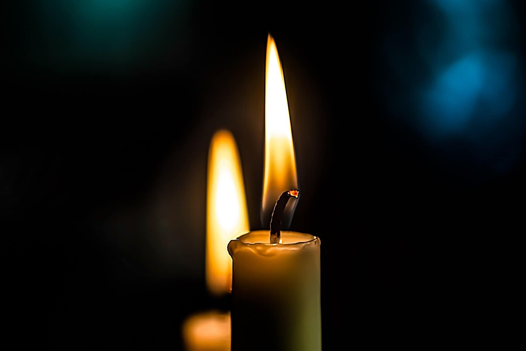 Two candles of staggered height are burning one behind the other in a dark space.
