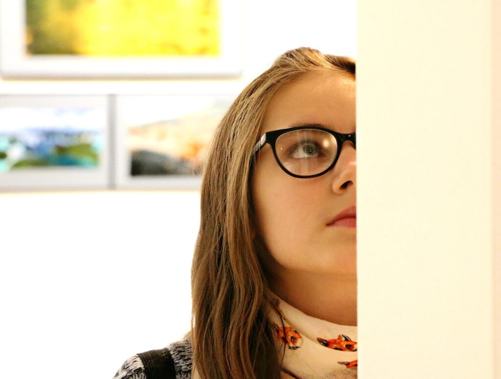 Half of a woman's face is blocked by a wall as she views a piece of art at a gallery.