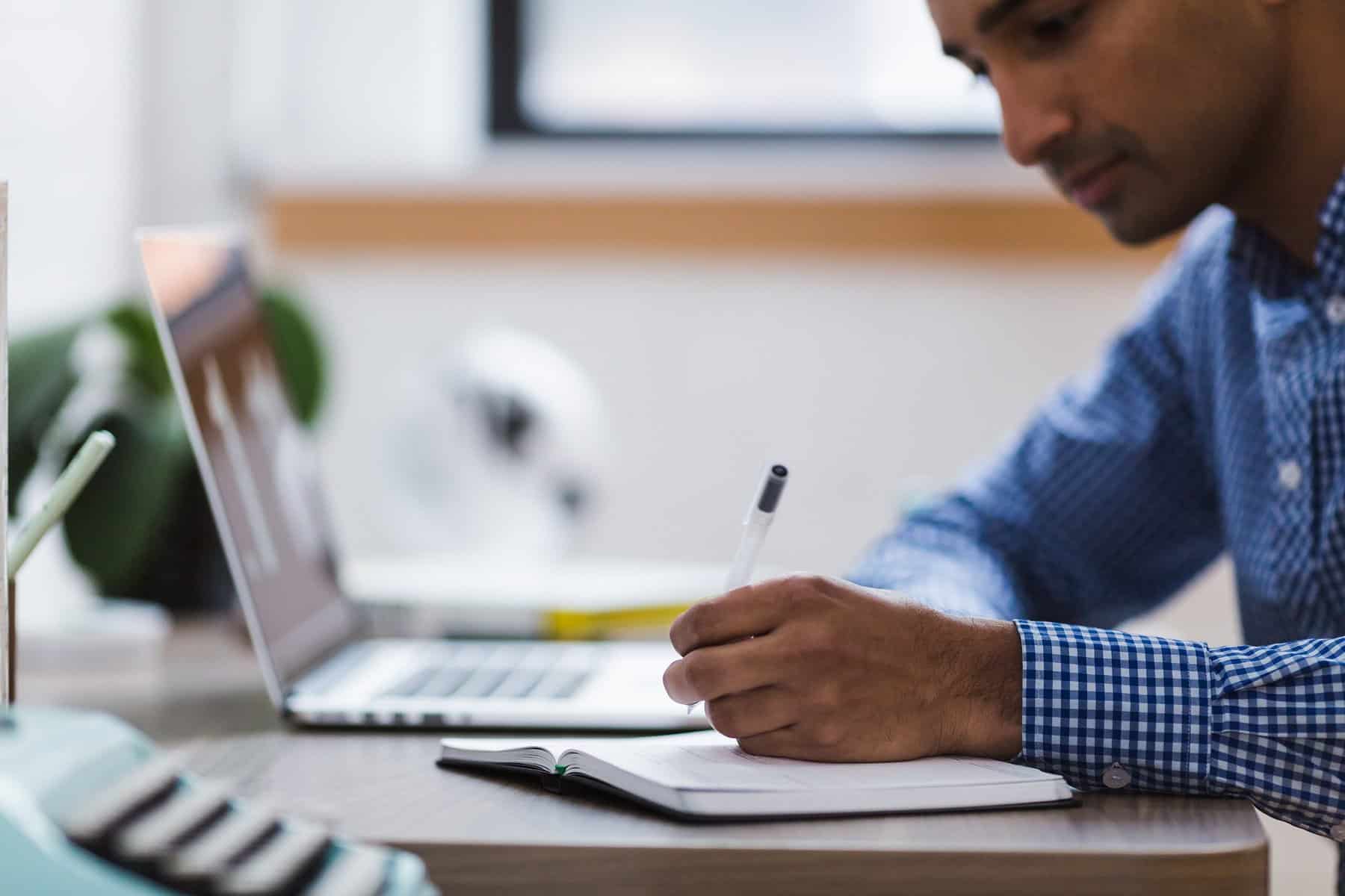 A man sits at desk with a computer and holds a pen over paper.
