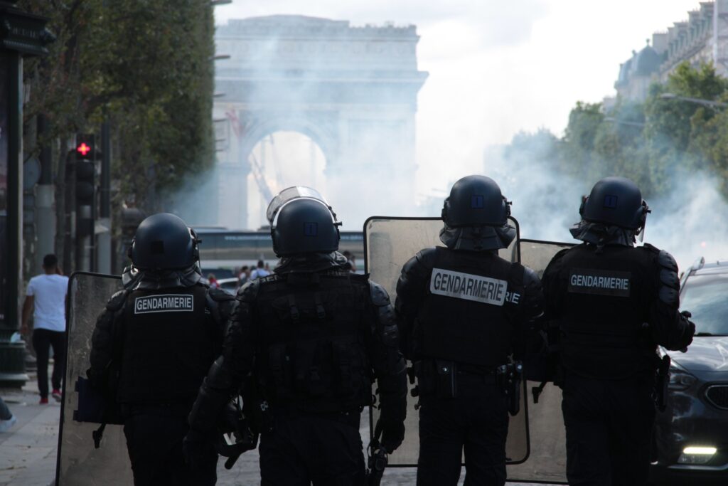 People in uniform hold riot shields and face a smoky road.