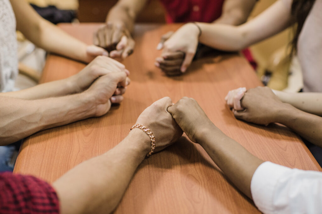 The arms of several people are shown around a small table holding the hands of their neighbors.