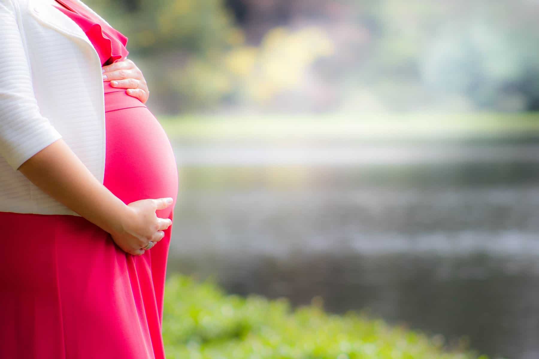 A woman's pregnant belly is shown with vegetation and a body of water behind her.