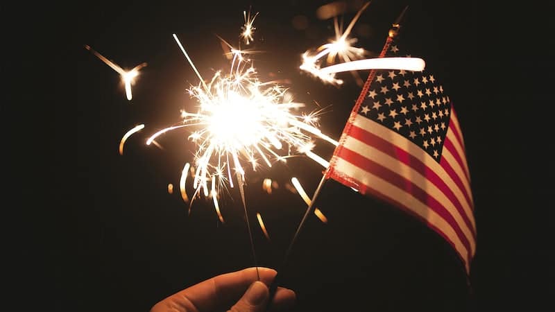 Faith and the Fourth of July A hand is holding a small American flag against a dark sky with a sparkler behind it.