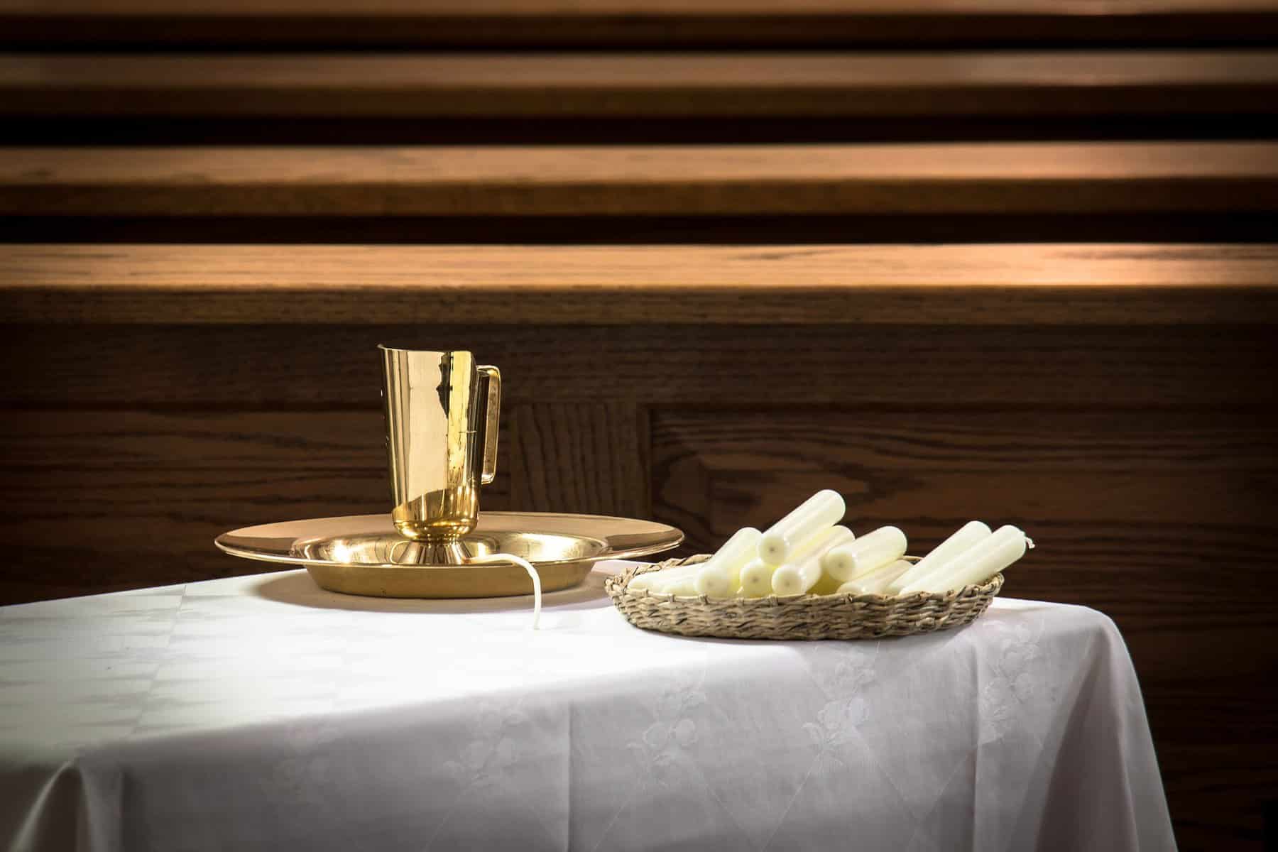 White candles sit in a basket behind church pews, with a gold chalice sitting in a gold plate beside them.