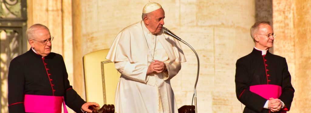Pope Francis appoints new patriarch of Lisbon after World Youth Day The Pope is seen dressed in white with two men on either side dressed in black with red sashes.