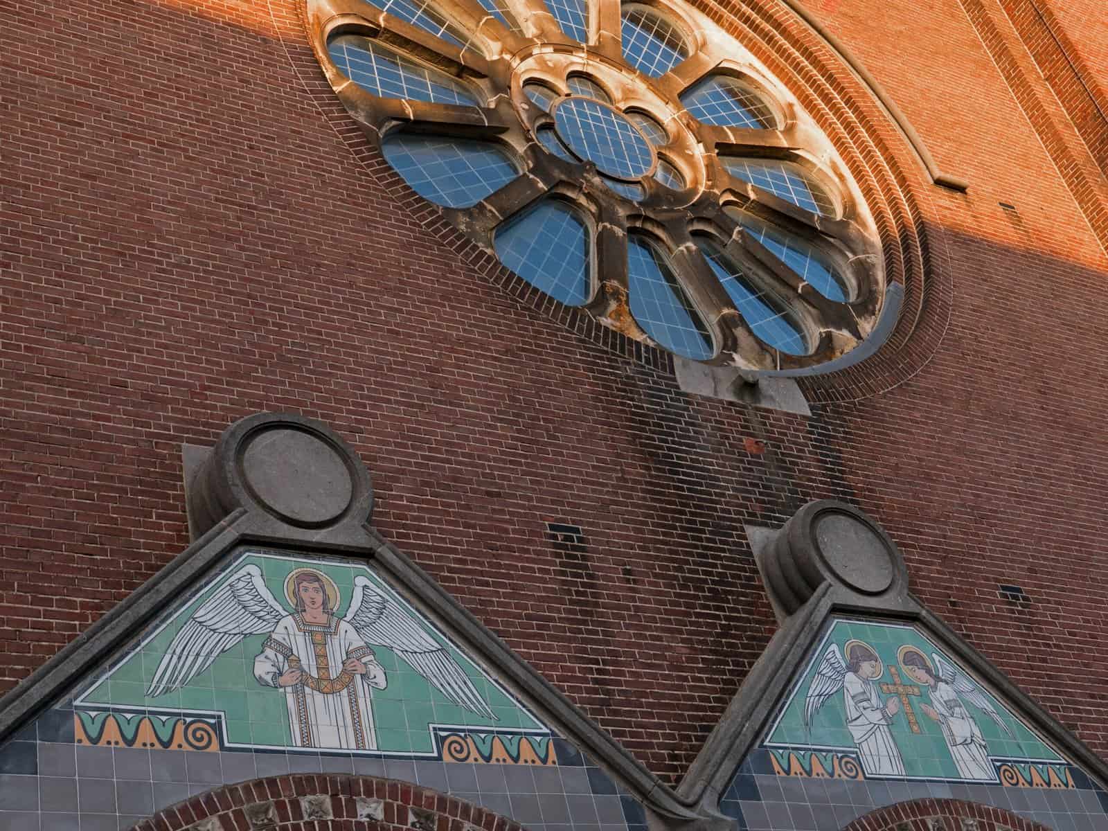 A historic red brick church facade in Amsterdam with tile work and a rose window are seen close up from below with camear angled up.