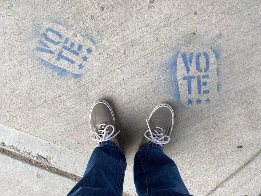 Maybe a second Trump term won’t be bad? The legs and feet of a person in blue jeans and canvas tennis shoes stands looking down at two blue paint stenciled "Vote" on concrete.