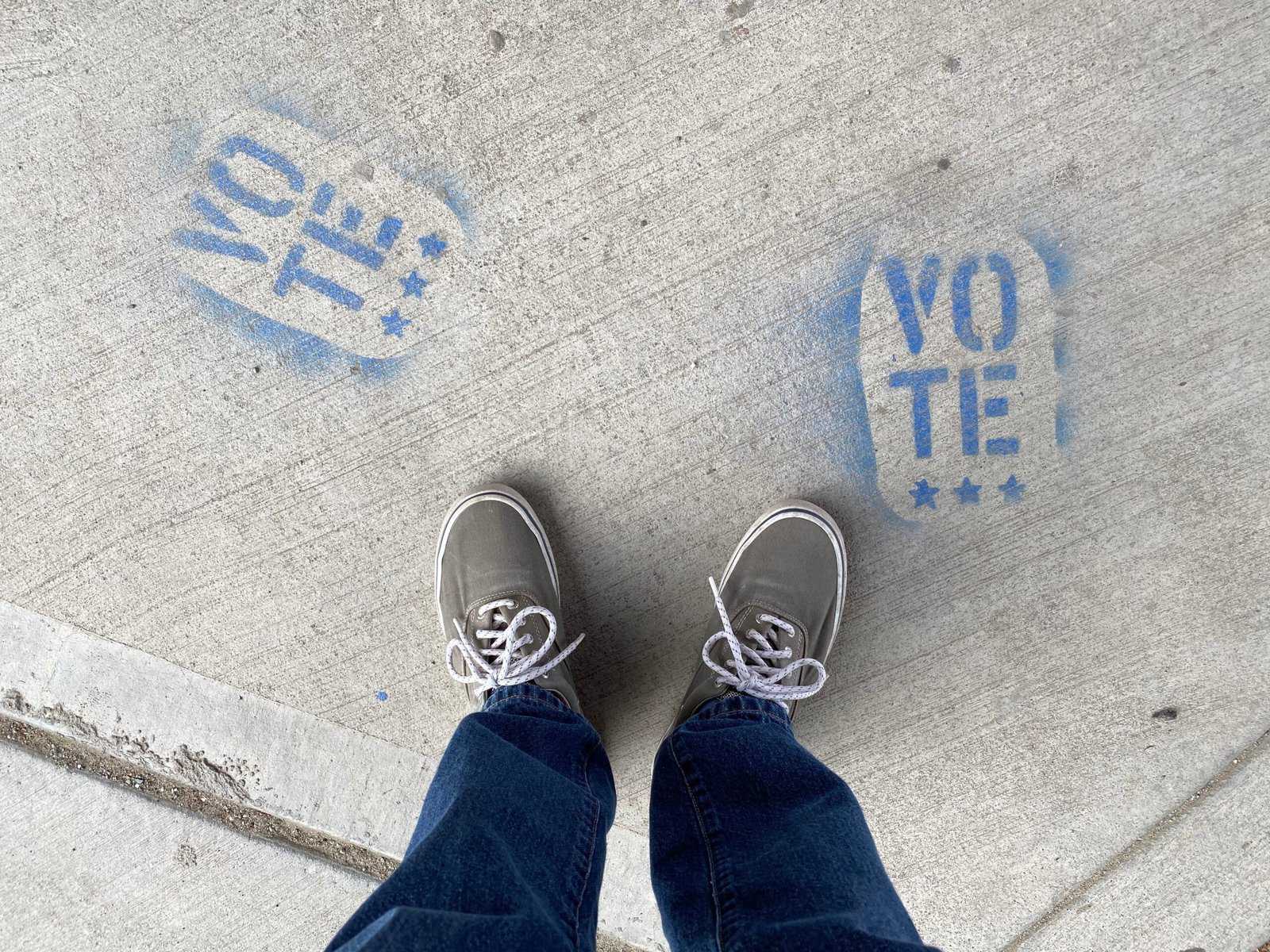 The legs and feet of a person in blue jeans and canvas tennis shoes stands looking down at two blue paint stenciled "Vote" on concrete.