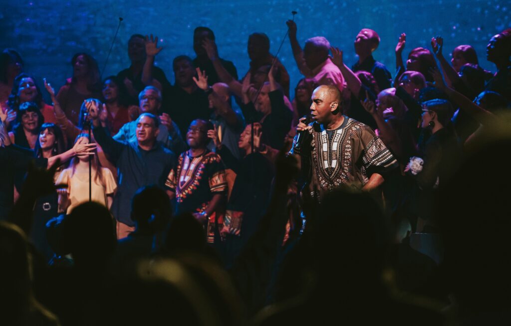 A group of Black and Caucasian people are in front of a church with stage lighting and some have their hands raised.