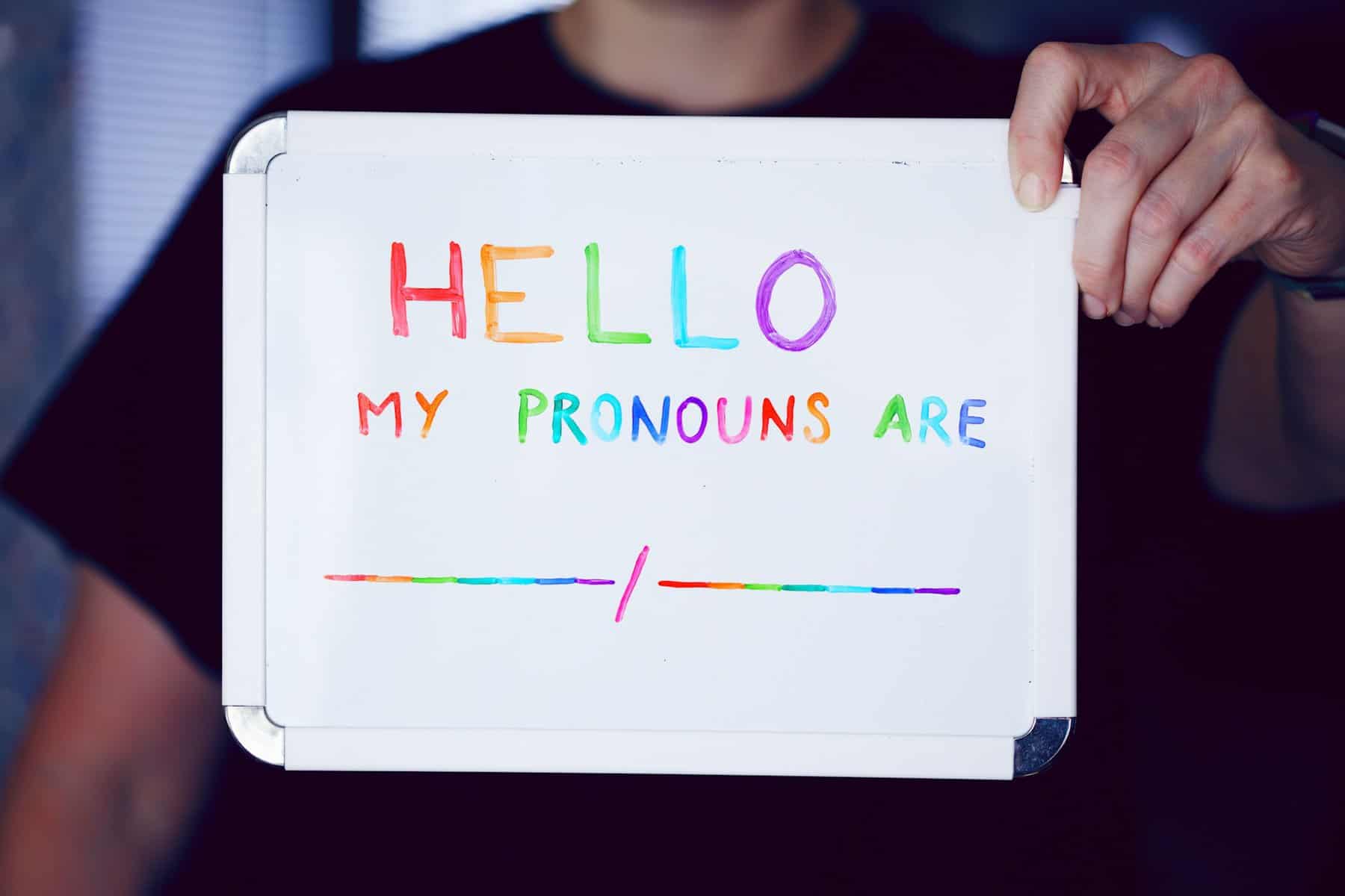 A person holds up a small white board with multi-colored words saying "Hello, my pronouns are..."