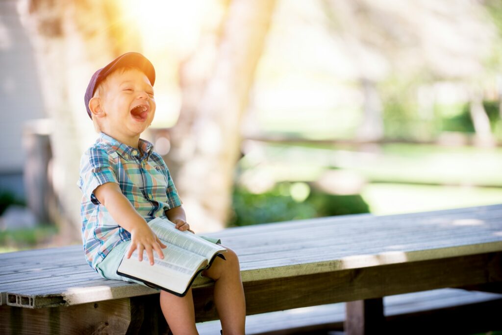Boy sitting on a bench in soft light and an open Bible on his lap.