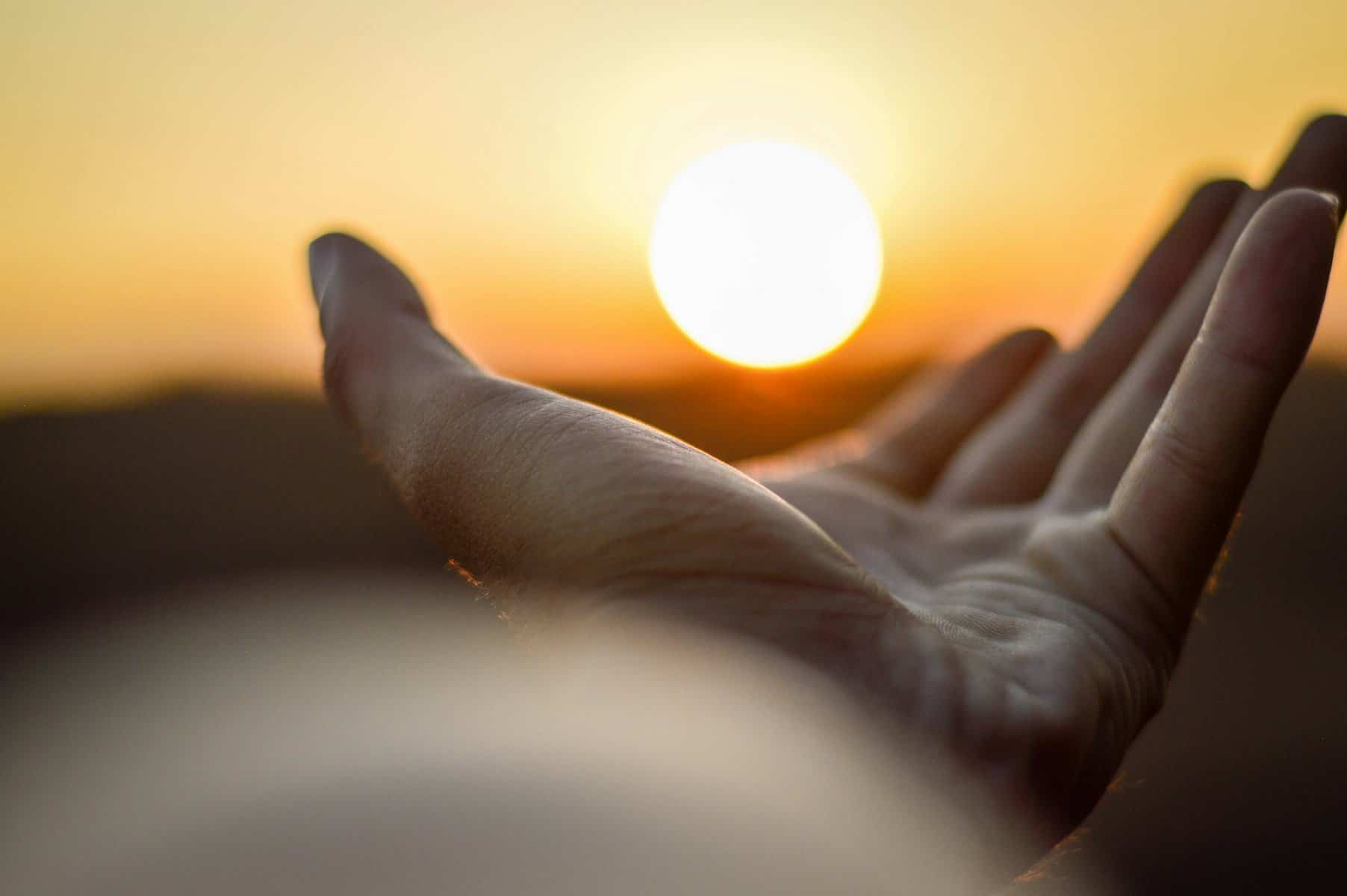 A man's hand reaches toward the setting sun as if holding it.