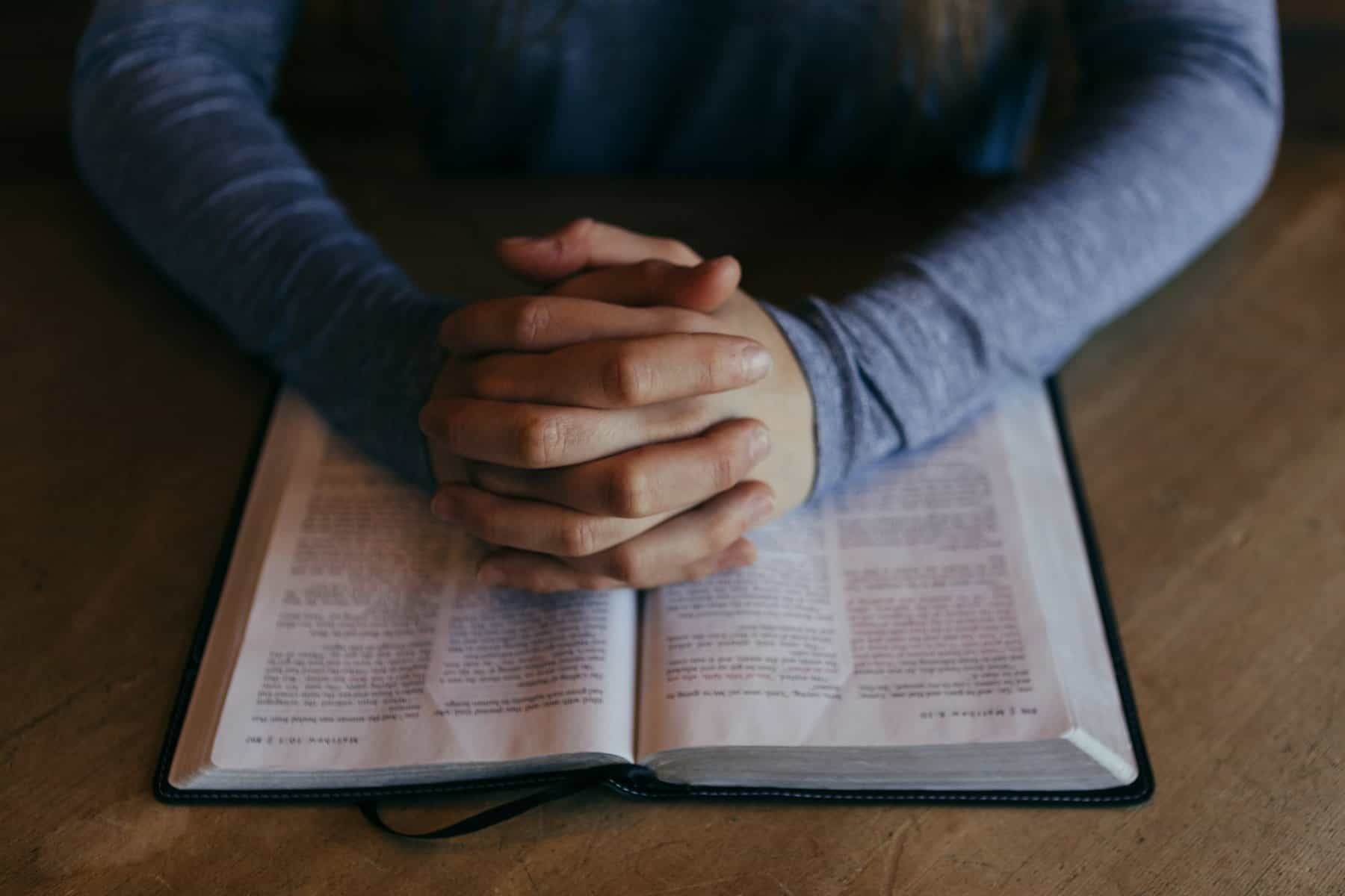 A man's hands are shown clasped and resting on a a Bible.