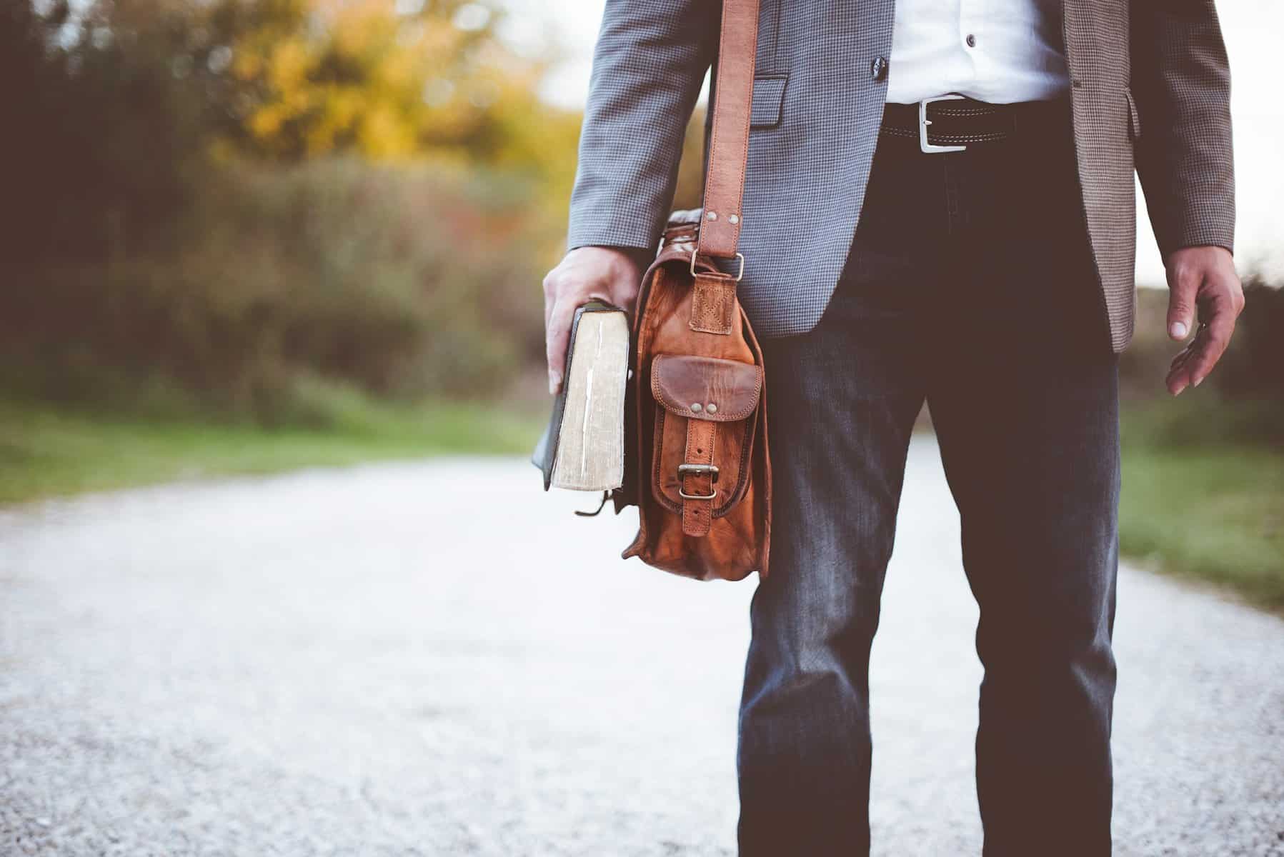 A man's torso is shown with a leather messenger bag on his shoulder, and holding a bible at his side.