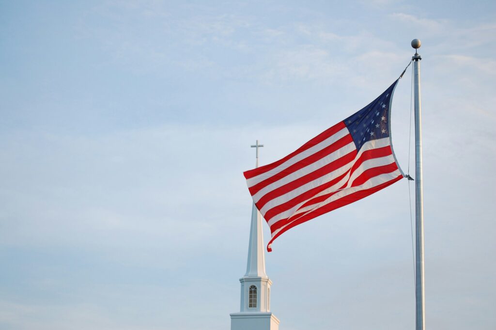 The growing rift in the Religious Right over Christian nationalism A church steeple topped with a cross behind a flying US flag.
