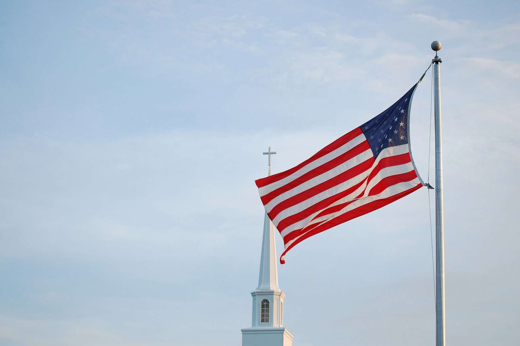 A church steeple topped with a cross behind a flying US flag.
