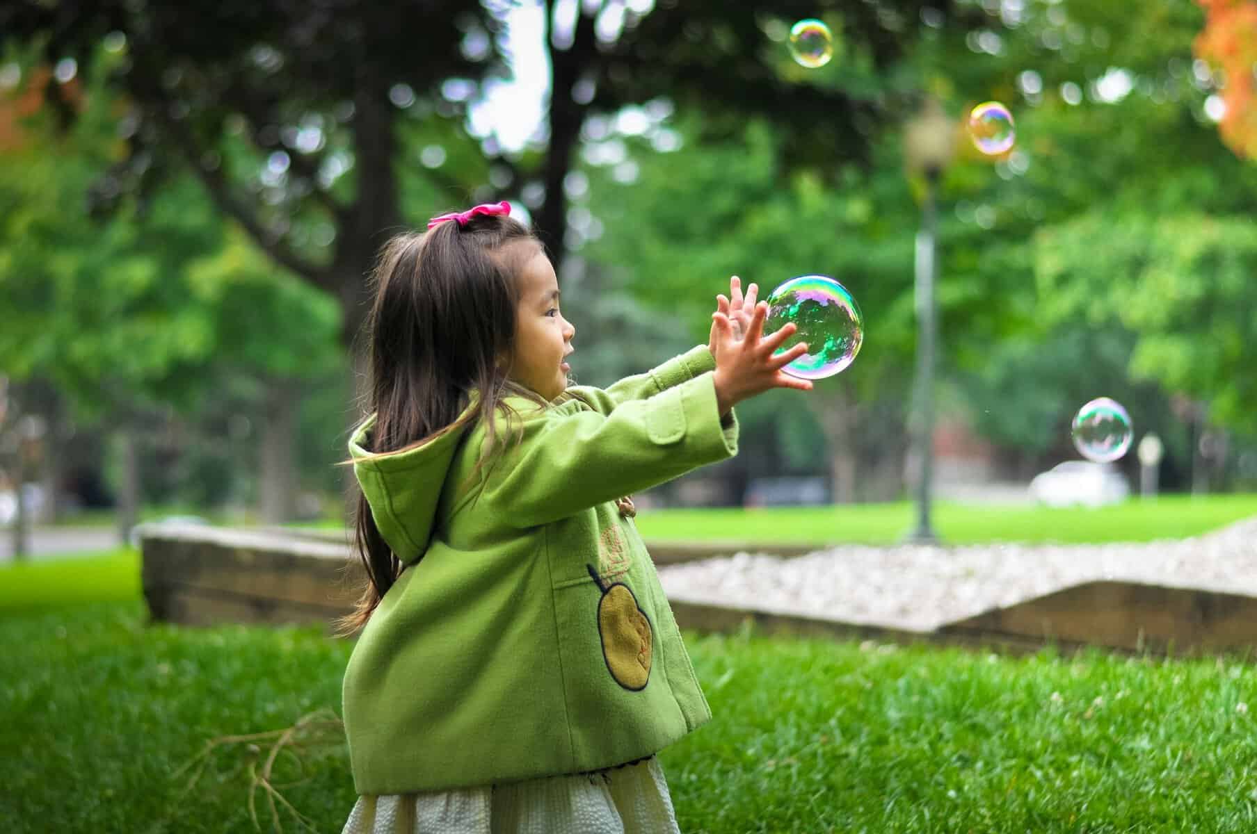 A toddler girl outside reaches for a large bubble with both hands.