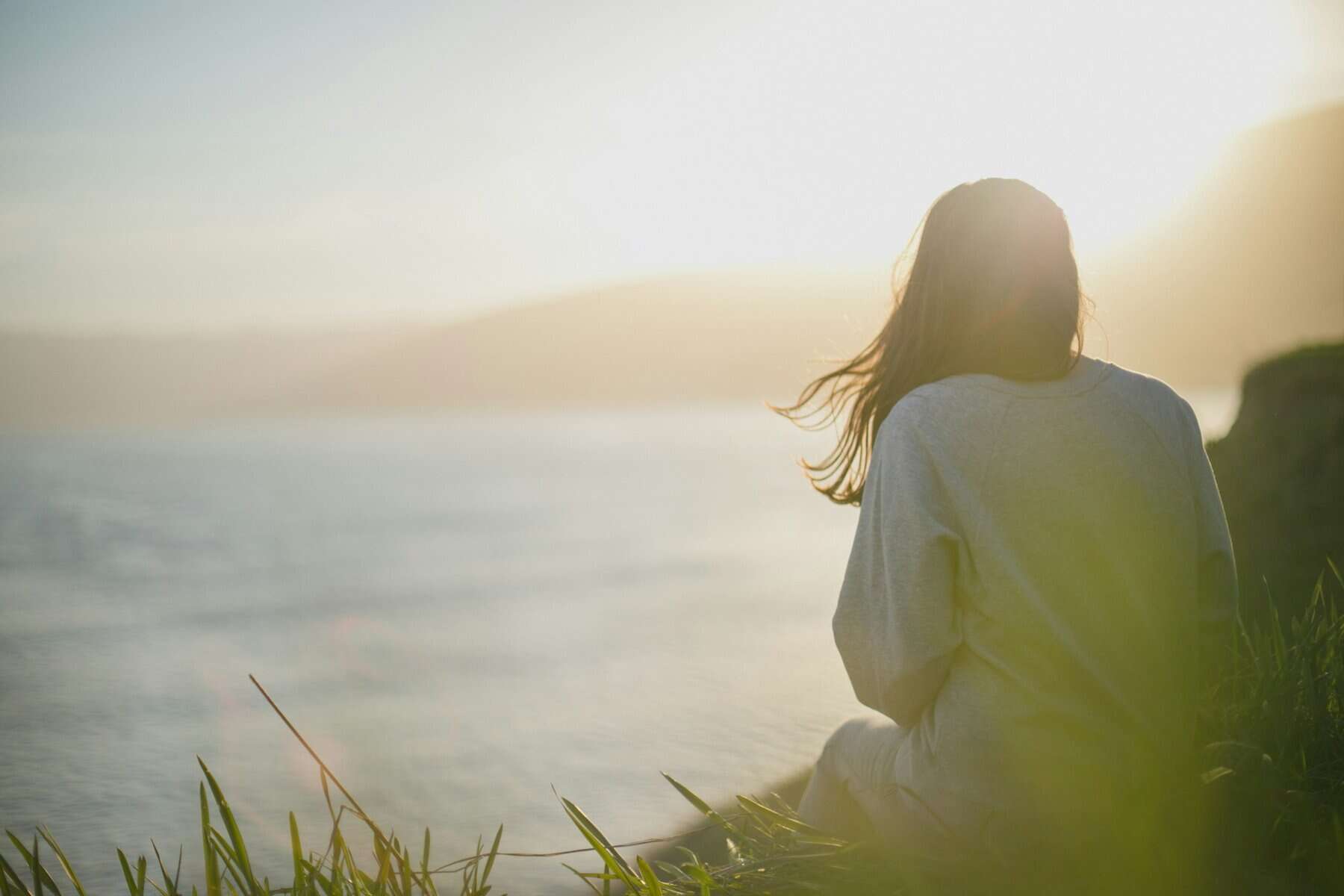 A woman sits by a body of water with her back to the camera.