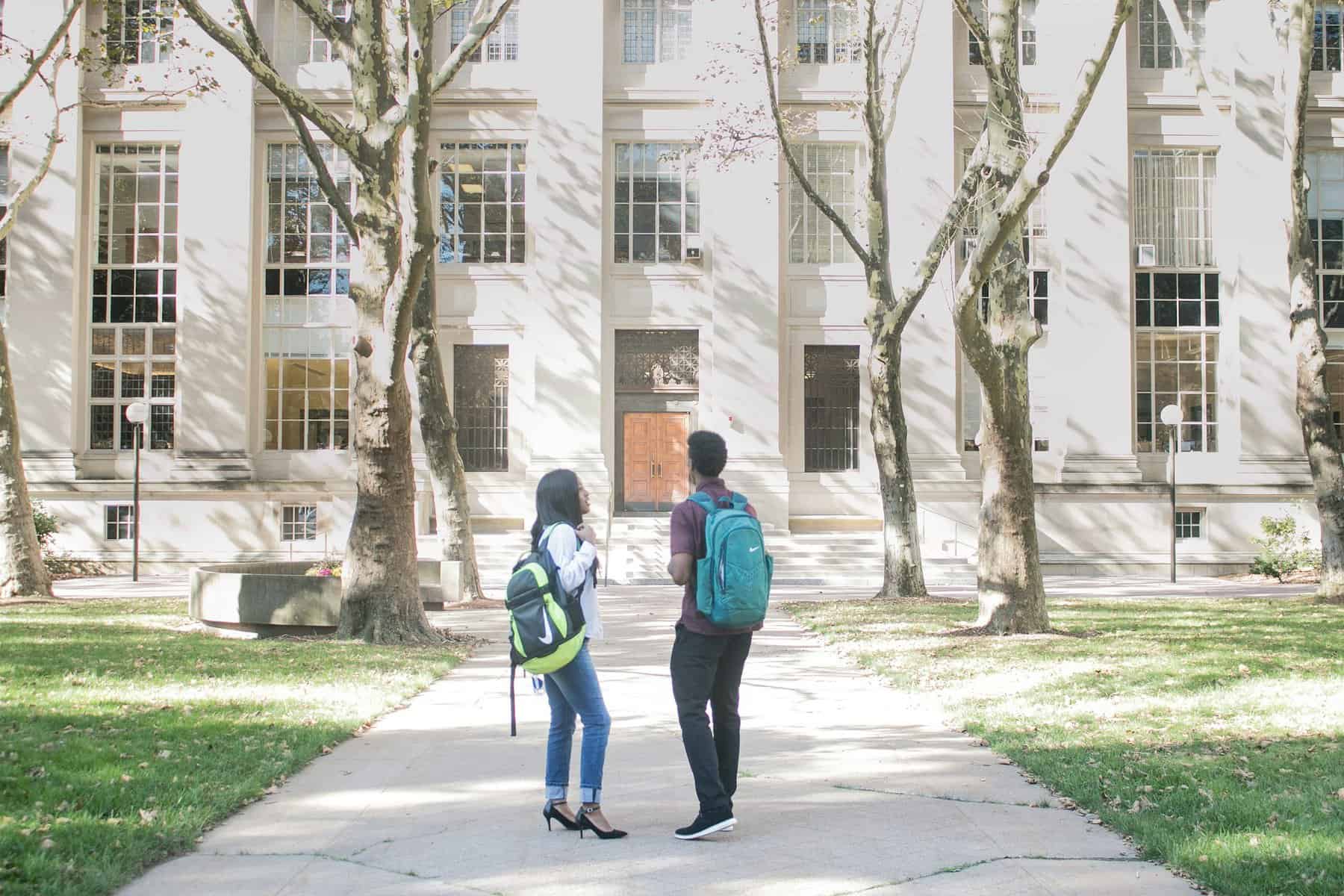 A man and a woman with backpacks stand in front of a large stone building.