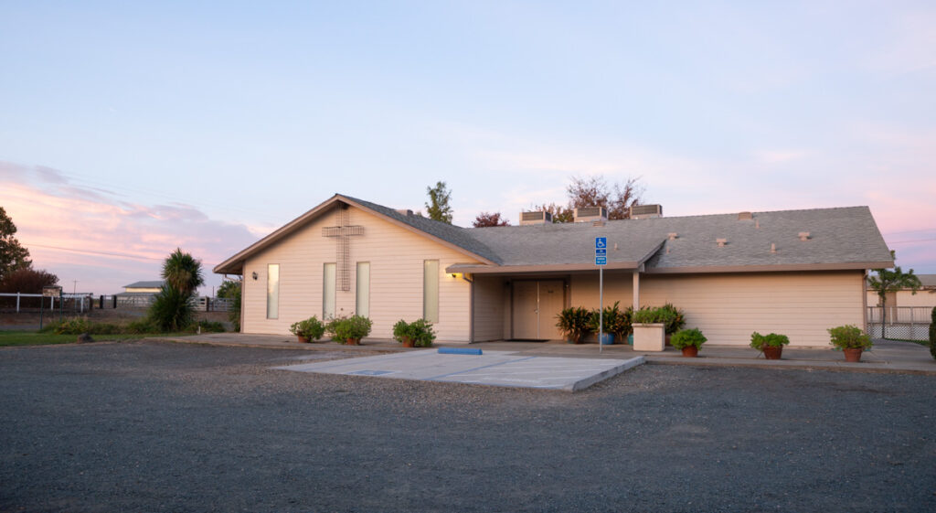 This image features a church building with a cross on the front, set in a gravel parking lot with potted plants and a serene sunset sky.