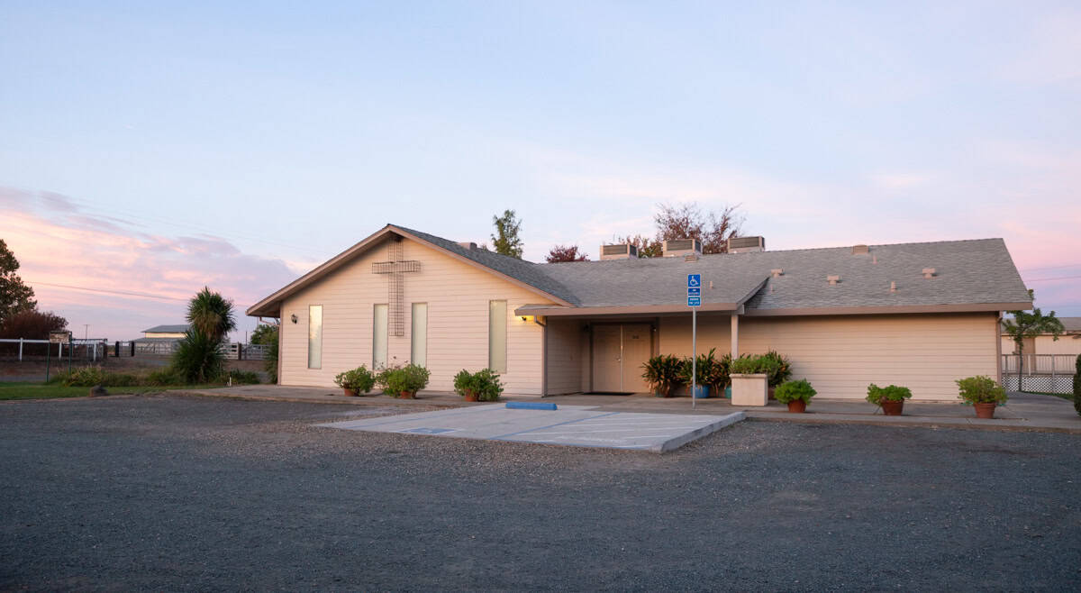 This image features a church building with a cross on the front, set in a gravel parking lot with potted plants and a serene sunset sky.