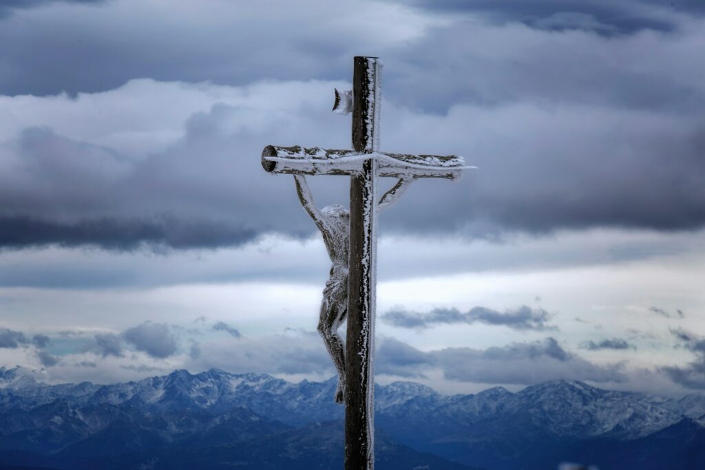 A wooden cross with a Christ figure covered in snow stands against a dramatic backdrop of snow-capped mountains.