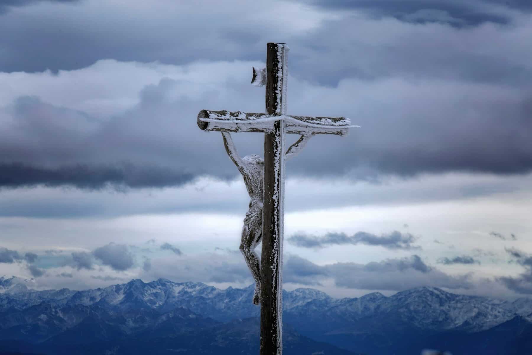 A wooden cross with a Christ figure covered in snow stands against a dramatic backdrop of snow-capped mountains.