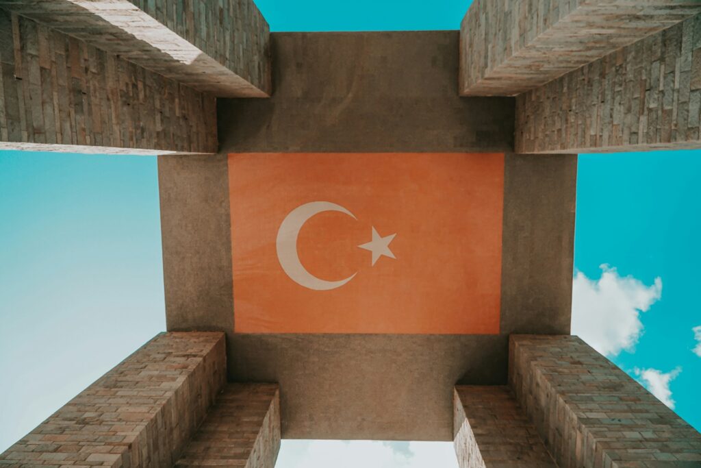 A striking image of the Turkish flag on a contemporary monument structure, showcasing national pride, faith, and cultural heritage, set against a vibrant blue sky with scattered clouds. Ideal for websites emphasizing Turkish identity, patriotism, or faith exploration.