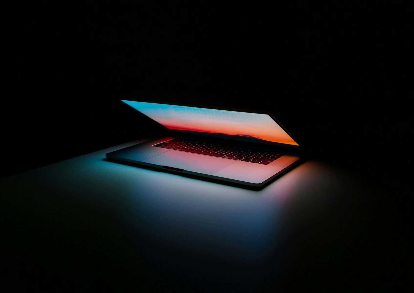 A half opened laptop in a dark room casting colorful light on the surface it's sitting on.