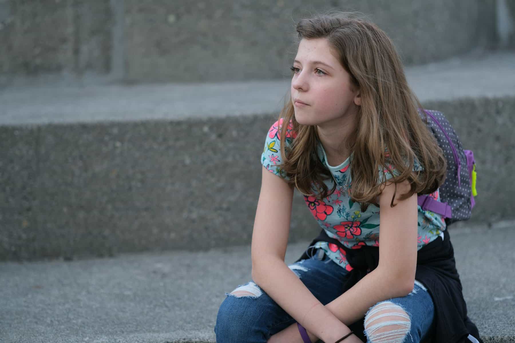 Young girl with long brown hair, dressed in a colorful floral t-shirt and ripped jeans, sitting on steps looking away and with a small backpack on her back.