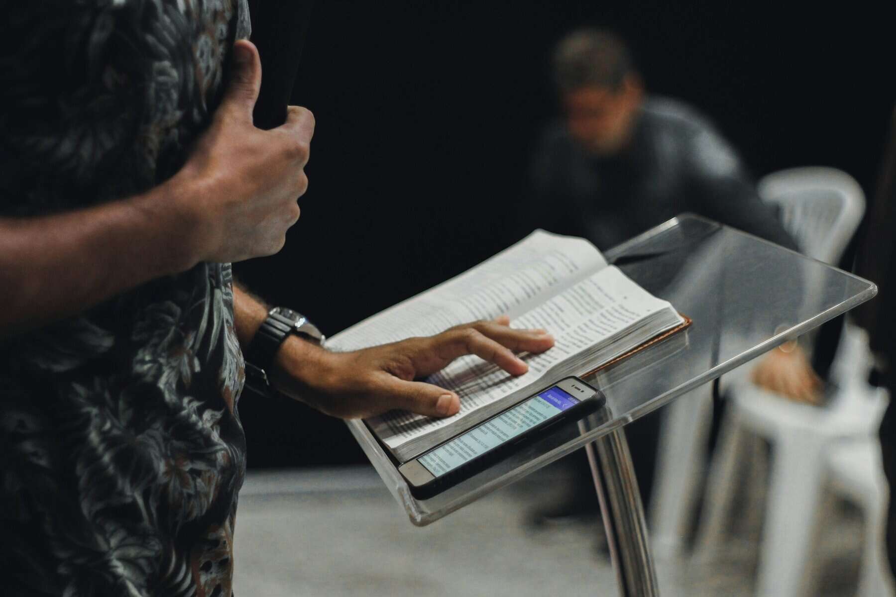 A man standing at a lectern with an open Bible and holding microphone.