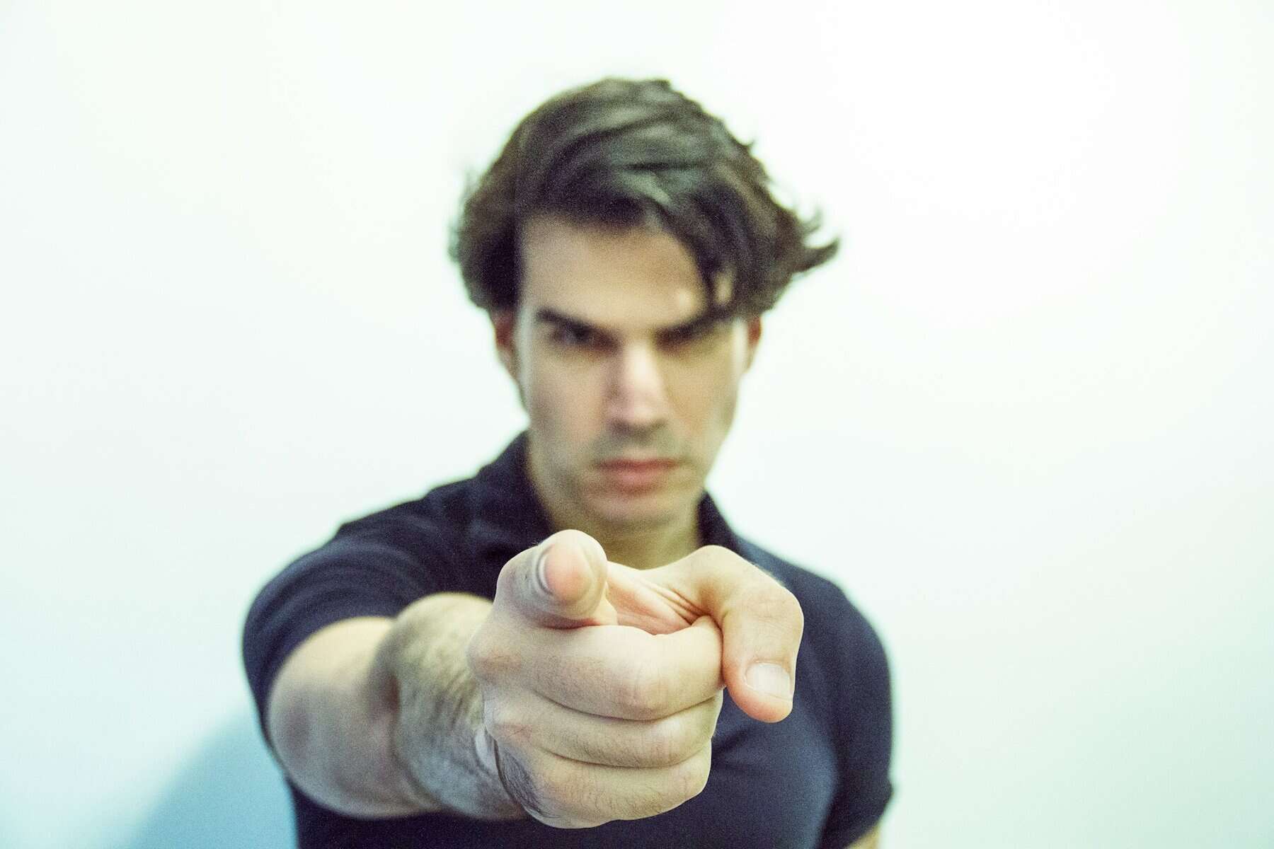 A man stands in front of a camera with a serious facial expression and index finger pointed at the camera and in focus.